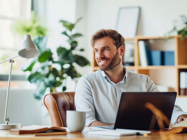 Mann arbeitet lächelnd im Homeoffice an einem Laptop – modernes, helles Arbeitsumfeld mit Pflanzen und Büromöbeln im Hintergrund.