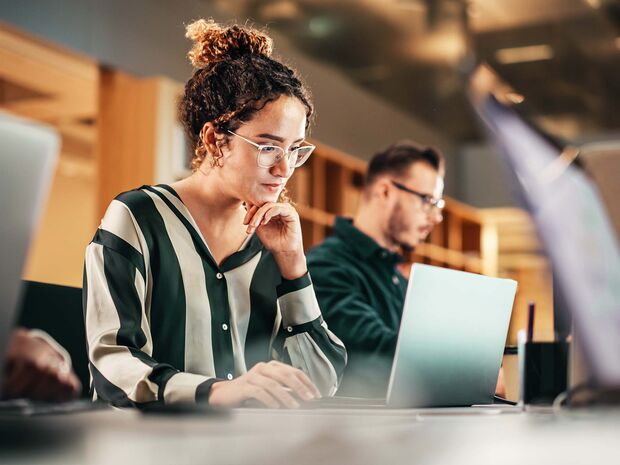 Frau sitzt am Tisch mit Laptop, im Hintergrund ein Mann mit Brille