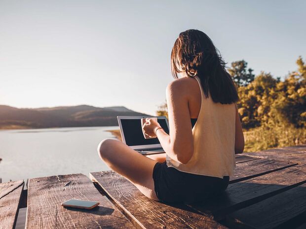 A woman sitting by a lake with her laptop and a cup of coffee