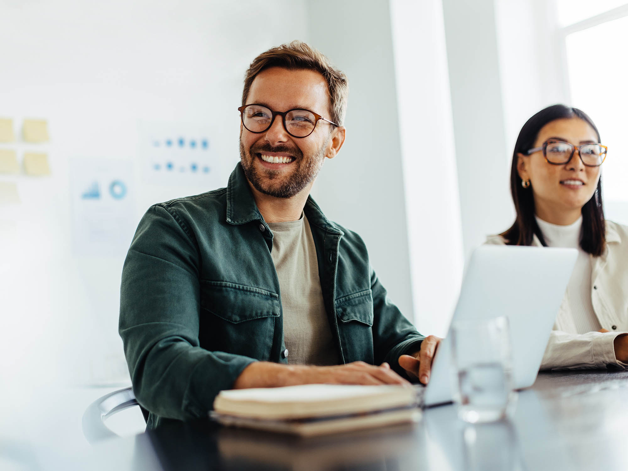 Un uomo e una donna siedono sorridenti a un tavolo con un computer portatile, un blocco note e un bicchiere d'acqua.